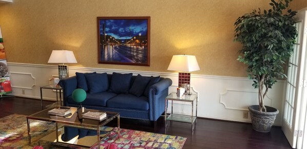 Uma sala de estar com um sofá azul, duas mesas laterais com abajures, uma mesa de centro de vidro, um tapete colorido, uma pintura de paisagem urbana emoldurada e uma planta em vaso.