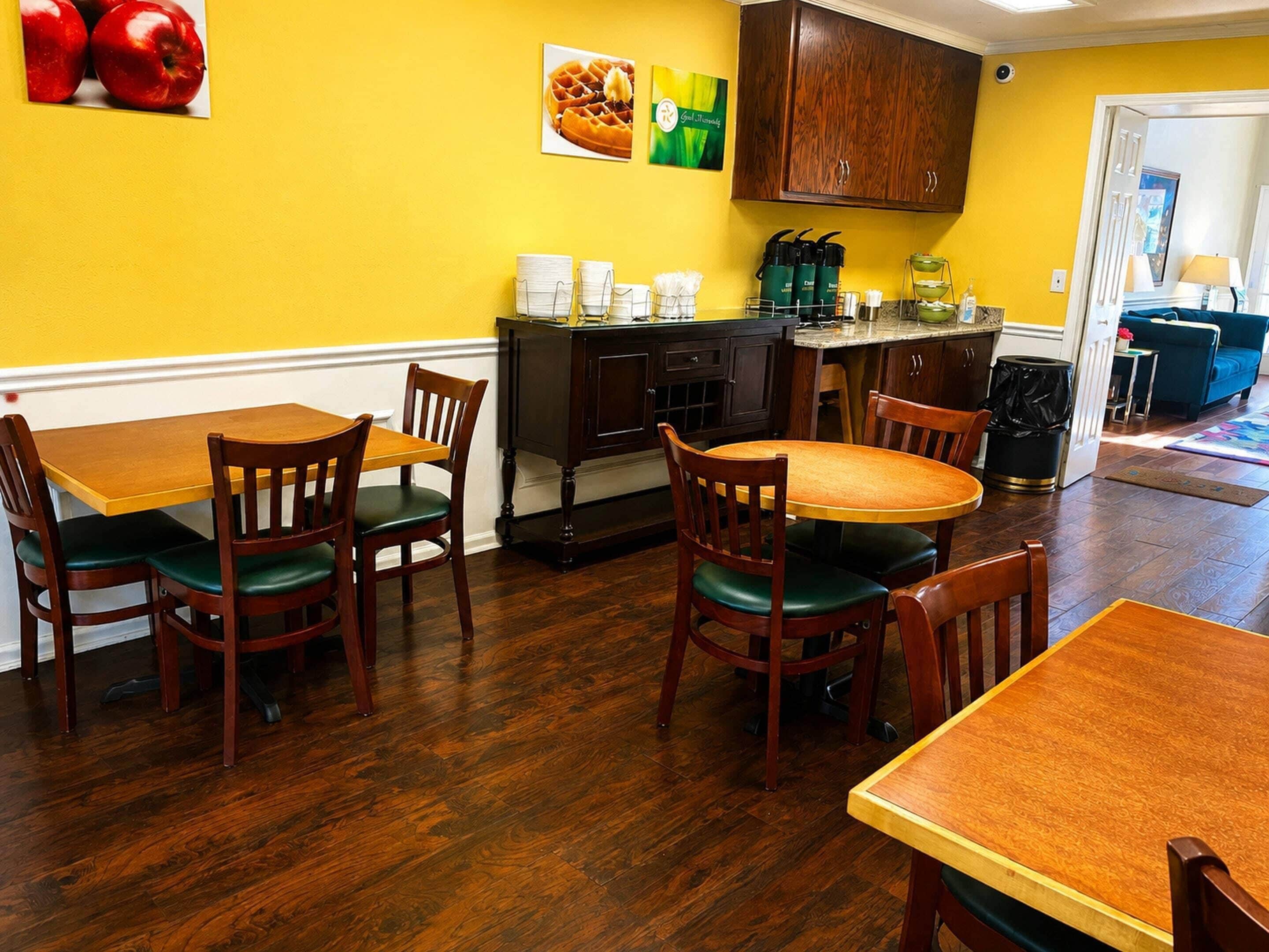 A dining area with wooden tables and chairs, a kitchen with dark cabinets, and a yellow wall.