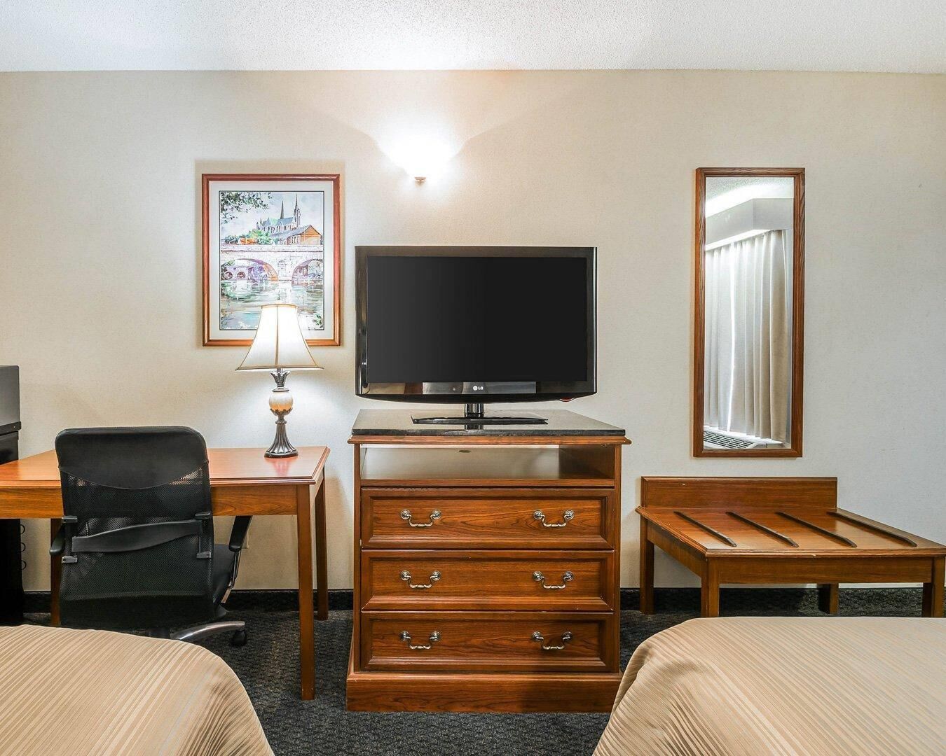 Una habitación de hotel con televisión de pantalla plana, un escritorio con lámpara, una silla, un aparador con cajones y dos camas.