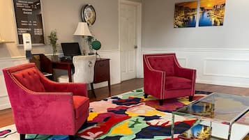 Um quarto com duas poltronas vermelhas estofadas, uma mesa de centro de vidro, um tapete colorido, uma escrivaninha com um laptop e um relógio fixado na parede.