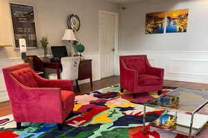 Um quarto com duas poltronas vermelhas estofadas, uma mesa de centro de vidro, um tapete colorido, uma escrivaninha com um laptop e um relógio fixado na parede.