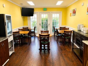 A dining area with wooden tables and chairs, a television, and a kitchen area with cabinets and a countertop.