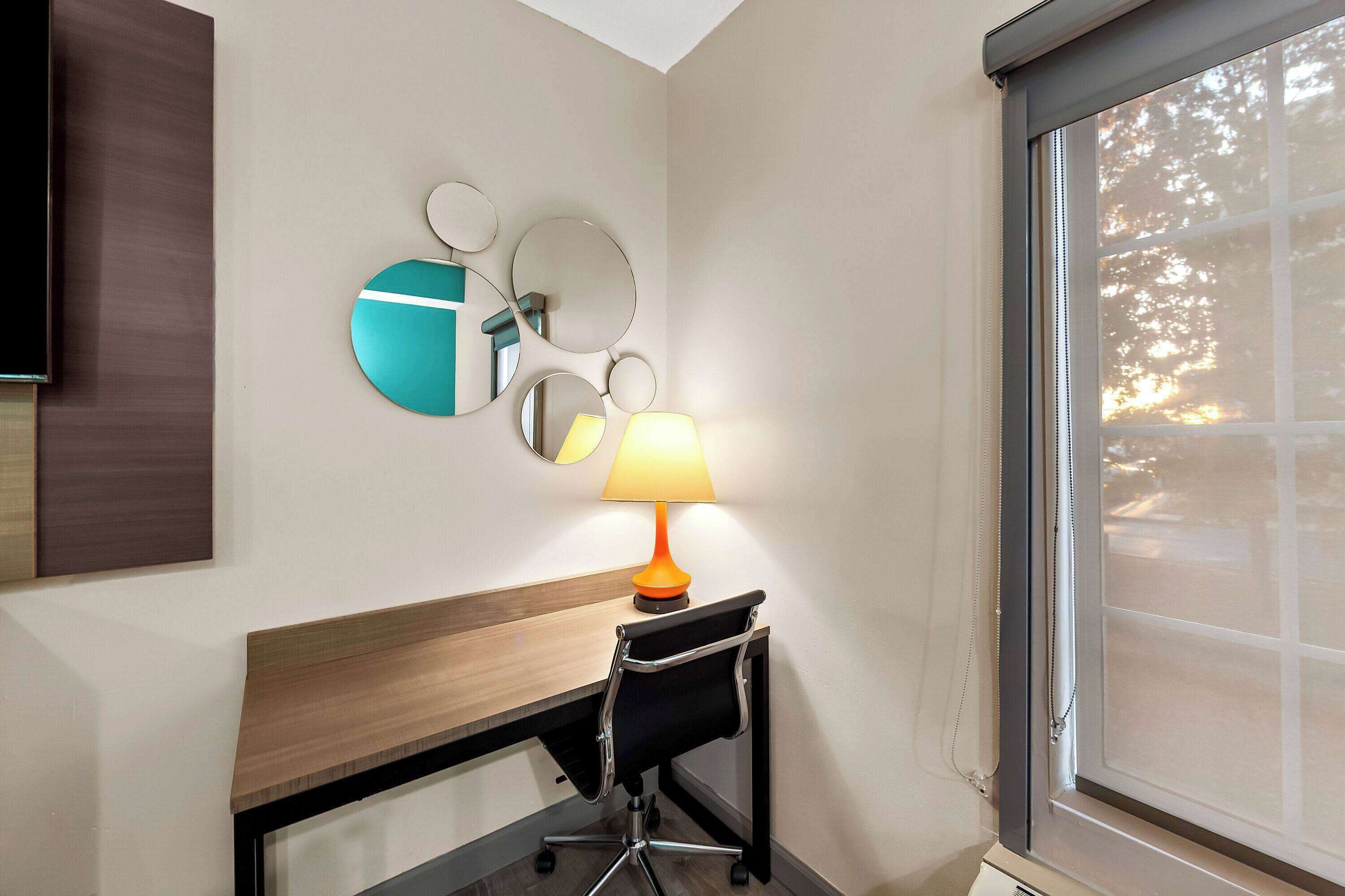 A hotel room with a wooden desk, a black office chair, a lamp, and a large window with a view of trees.