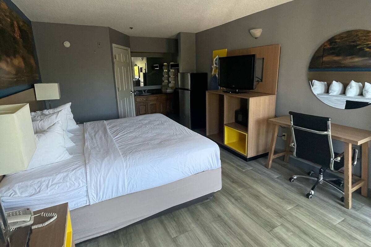 Une chambre d’hôtel comprenant un lit, un bureau avec une chaise, une télévision, un petit coin cuisine et une salle de bain visible à travers une porte ouverte.