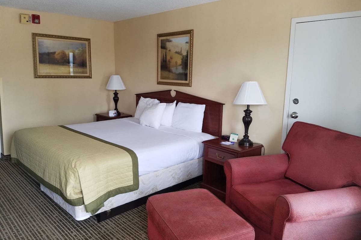 Une chambre d’hôtel avec un lit, deux tables de chevet avec des lampes, un fauteuil rouge et deux tableaux encadrés au mur.