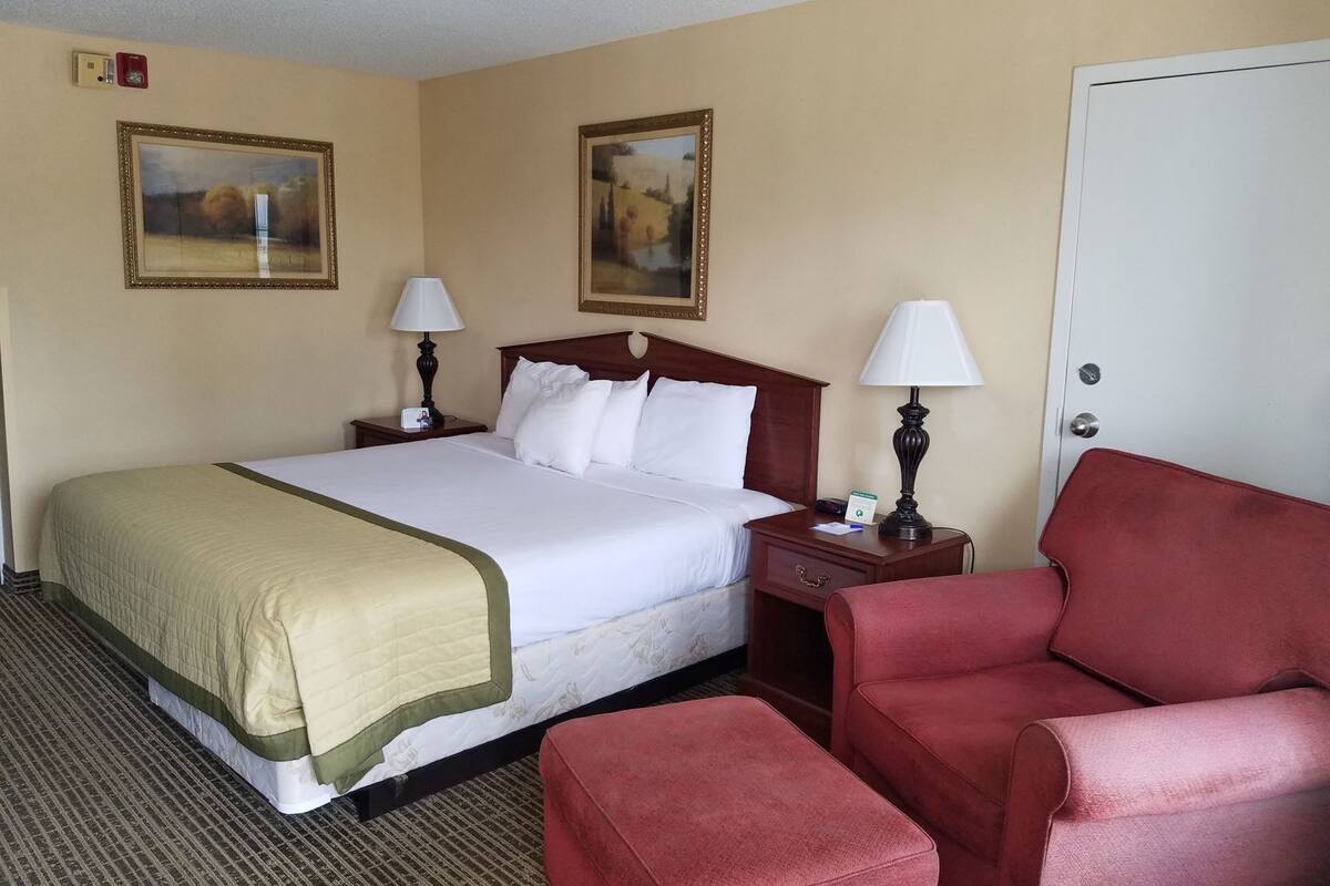 Une chambre d’hôtel avec un lit, deux tables de chevet avec des lampes, un fauteuil rouge et deux tableaux encadrés au mur.