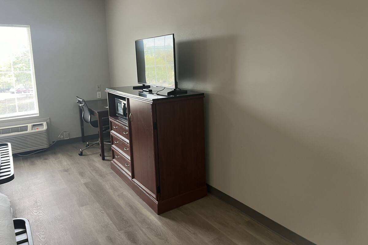 A small, well-lit room with a wooden desk, a chair, a television, and a window with blinds.