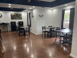 Comedor con mesas y sillas de madera, área de cocina con refrigerador y tostadora, y un reloj en la pared.