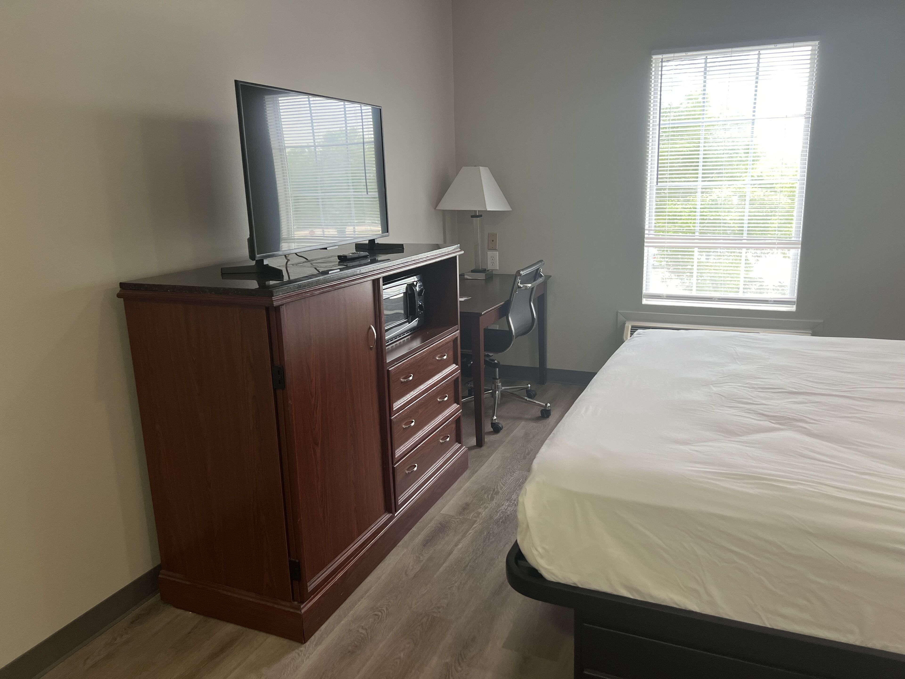 Une chambre à coucher comprenant un lit, une commode en bois, un bureau avec une chaise, un téléviseur et une fenêtre avec des stores.