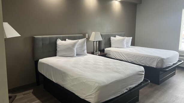 A hotel room with two beds, a nightstand with a lamp, and a window with blinds.