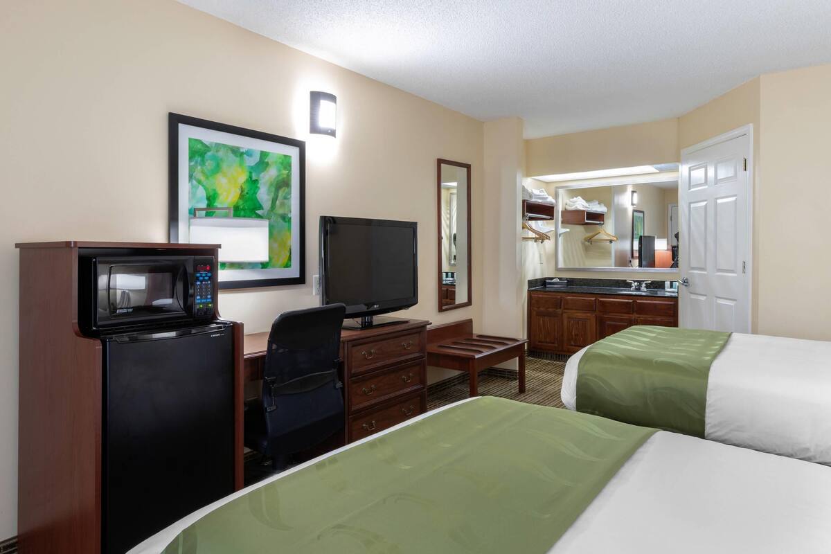 Quarto de hotel com duas camas, uma escrivaninha com micro-ondas, televisão e uma cozinha compacta ao fundo.
