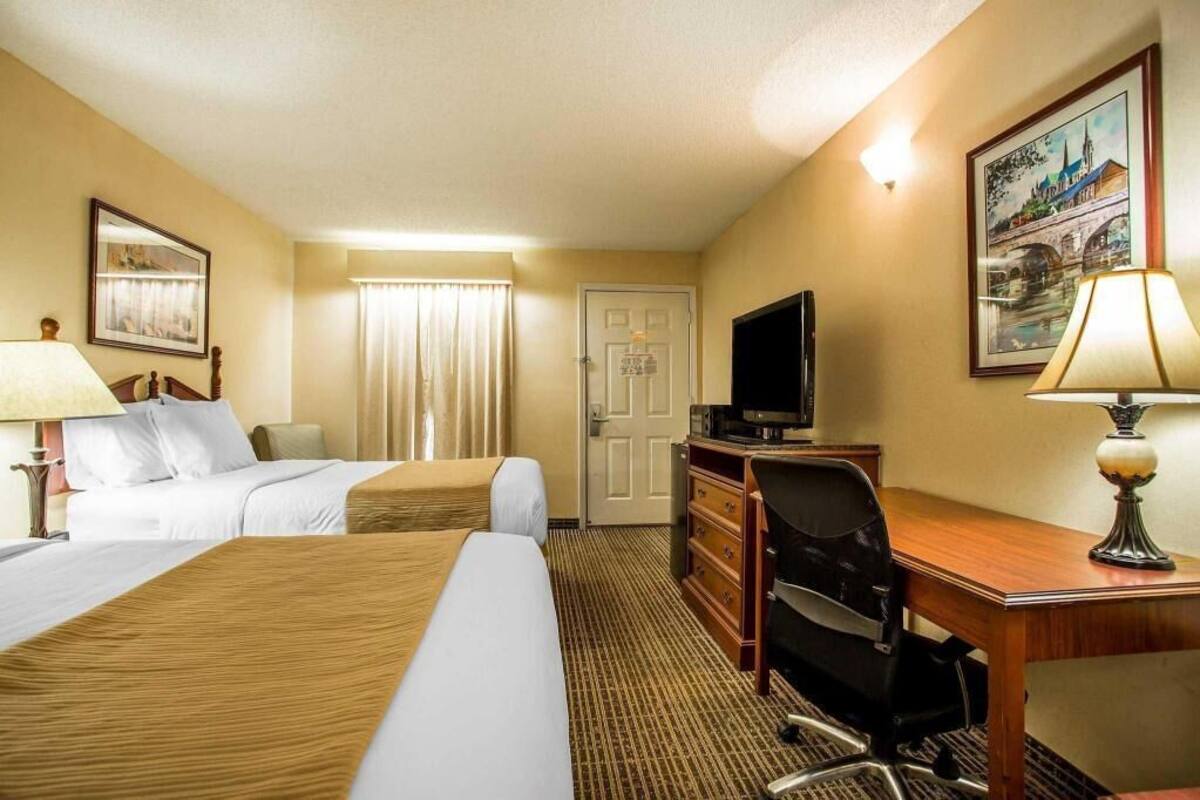 Quarto de hotel com duas camas, uma escrivaninha, uma cadeira, uma televisão e um abajur.