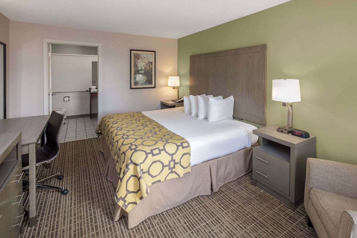 A hotel room with a bed, desk, chair, and a nightstand with a lamp.