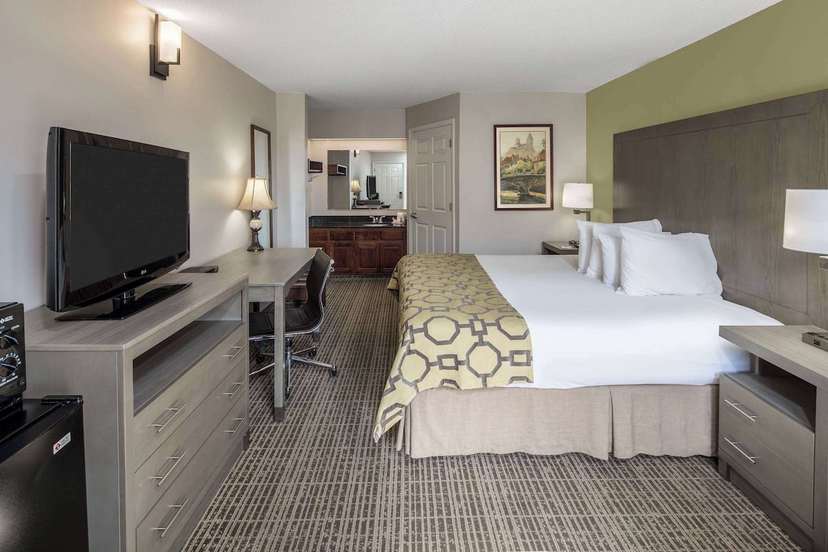 A hotel room with a bed, a desk with a television, a chair, and a kitchenette in the background.