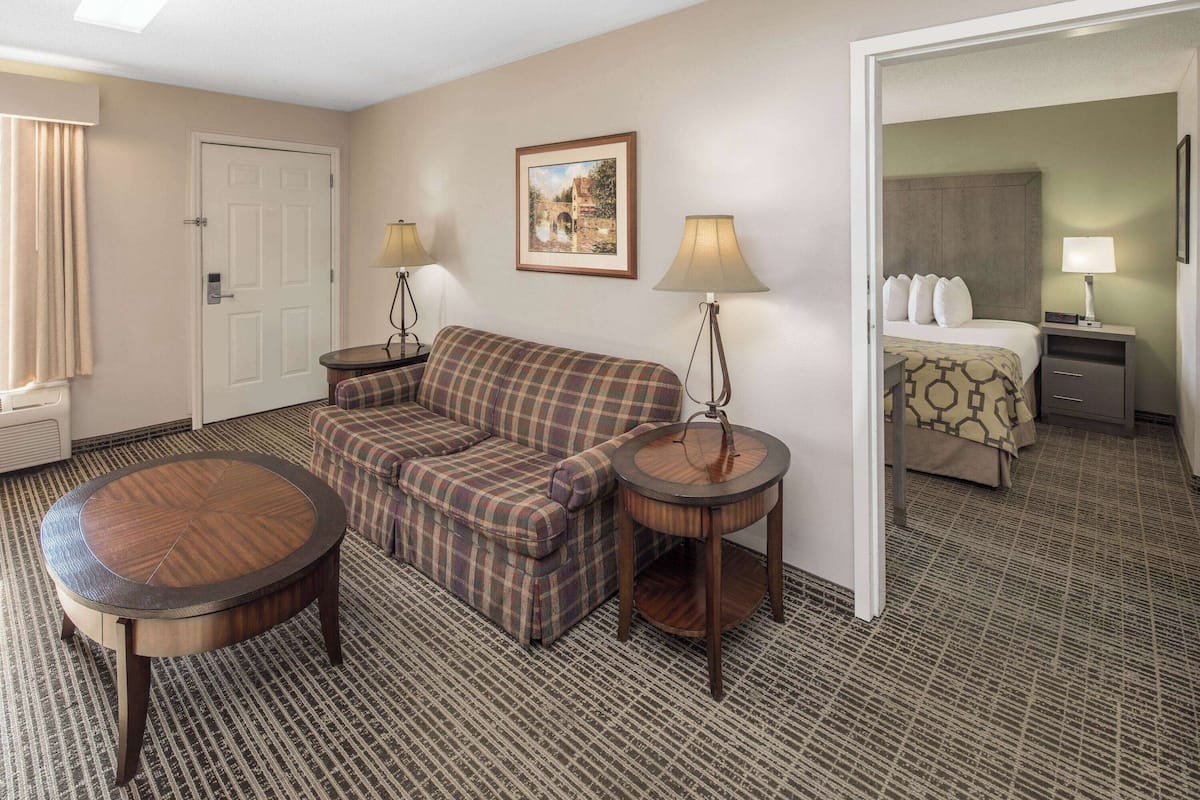 A hotel room with a plaid sofa, a round wooden table, two bedside lamps, a framed picture on the wall, and a bed with patterned bedding.