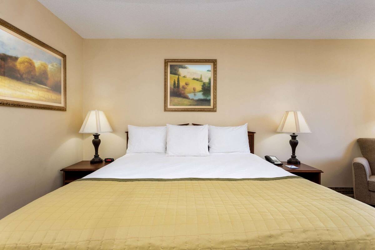 Habitación de hotel con una cama grande, dos mesitas de noche con lámparas, una silla y dos cuadros en las paredes.