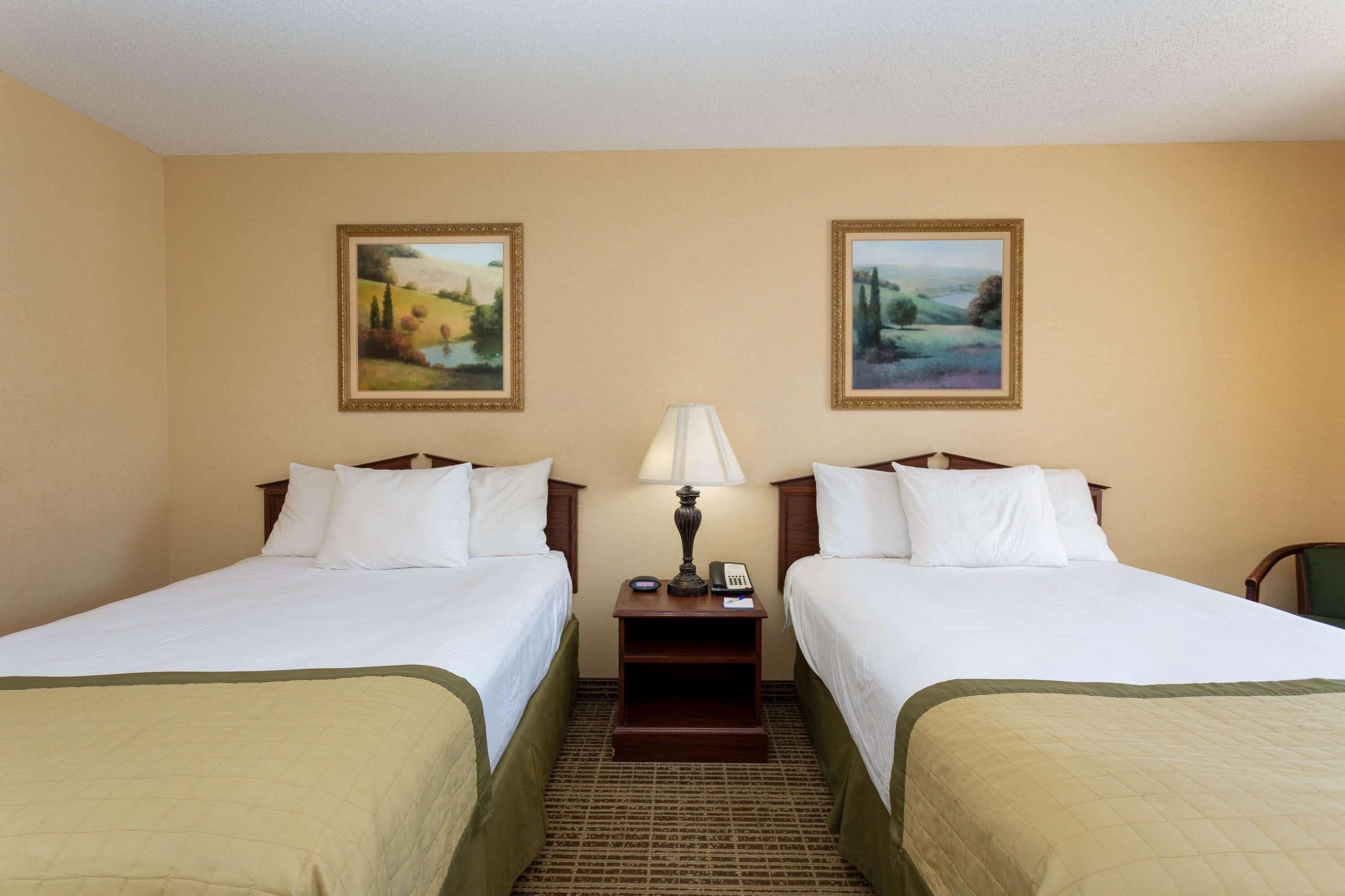 A hotel room with two beds, a nightstand with a lamp, and two framed landscape paintings on the wall.