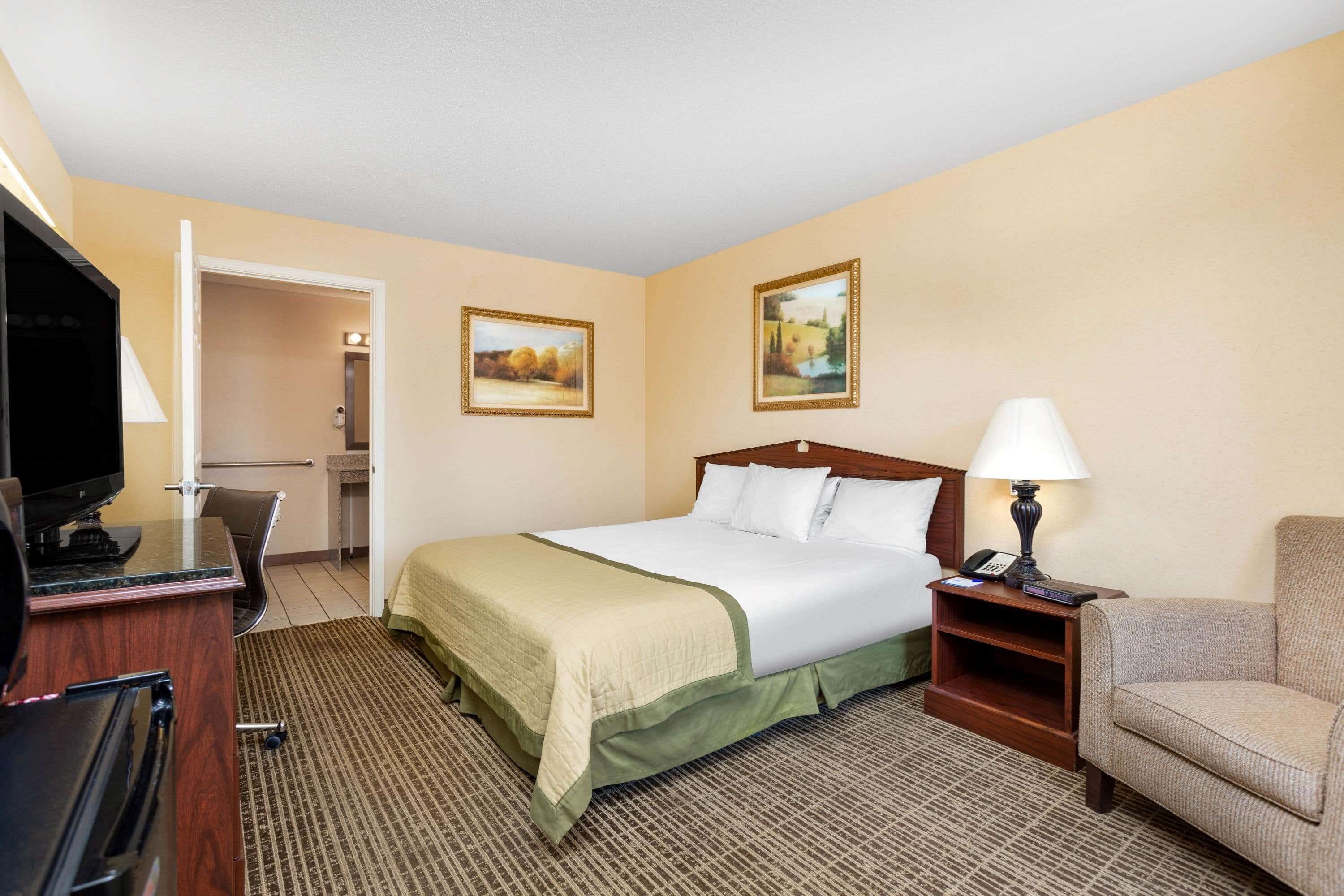 A hotel room with a bed, a desk with a computer, a chair, a TV, and two paintings on the wall.