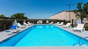 Una piscina con sillas de descanso y una silla para socorrista, rodeada por una valla y un edificio.