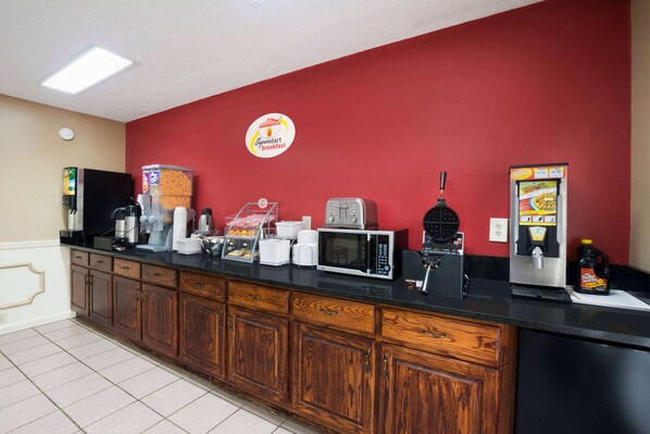 A breakfast bar with a red wall, a coffee machine, a microwave, a toaster, and a beverage dispenser.