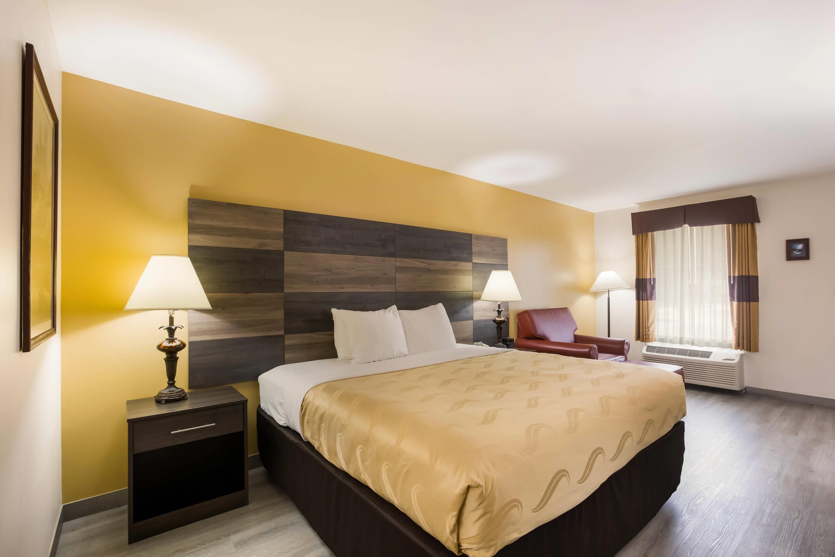 Habitación de hotel con una cama grande, dos mesitas de noche con lámparas, un sillón rojo, una ventana con cortinas y un aire acondicionado instalado en la pared.