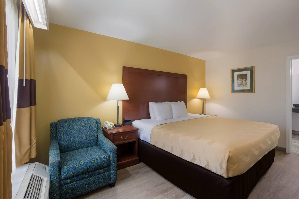 Ein Hotelzimmer mit einem Bett, zwei Nachttischlampen, einem Sessel, einem Nachttisch und einem an der Wand hängenden Bild.