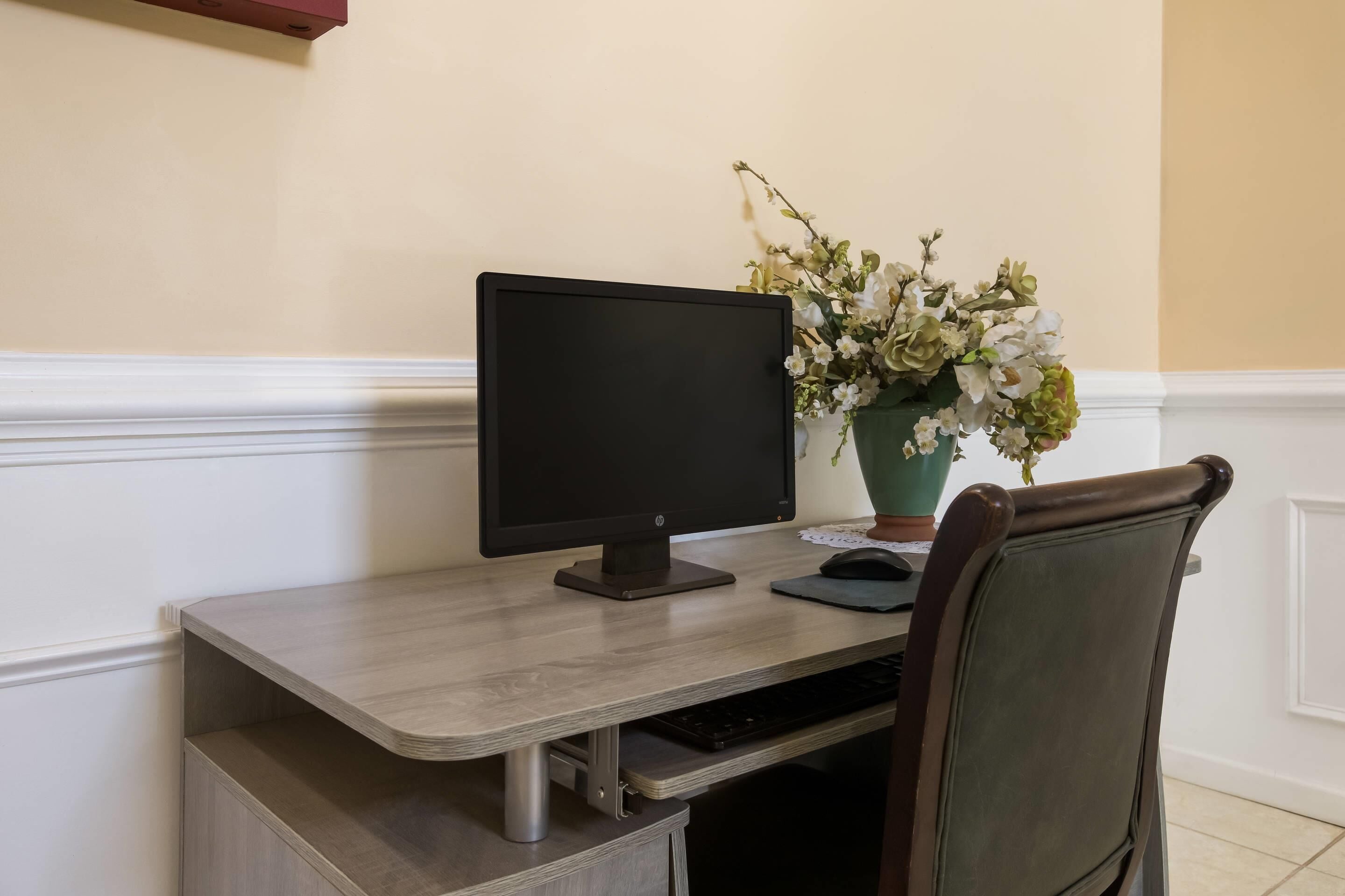 A desk with a computer monitor, keyboard, and a mouse. There is a chair with a cushioned backrest. A vase with flowers is placed on the desk.