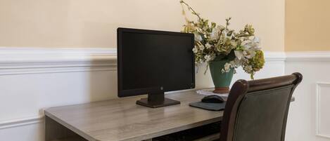 A desk with a computer monitor, keyboard, and a mouse. There is a chair with a cushioned backrest. A vase with flowers is placed on the desk.
