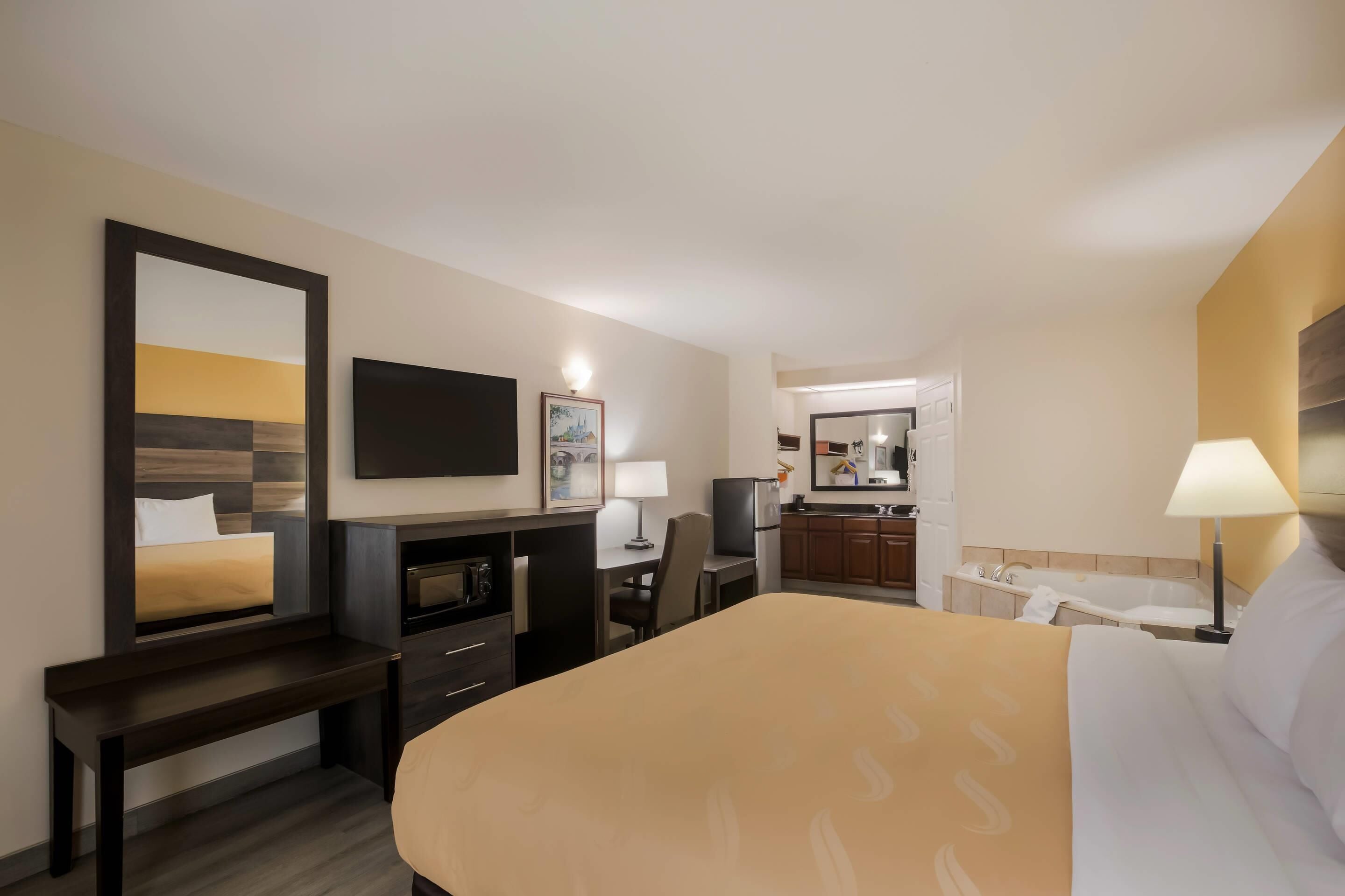 Ein Hotelzimmer mit Bett, Schreibtisch, Stuhl, Fernseher, Spiegel, Lampe und einer Küchenzeile.