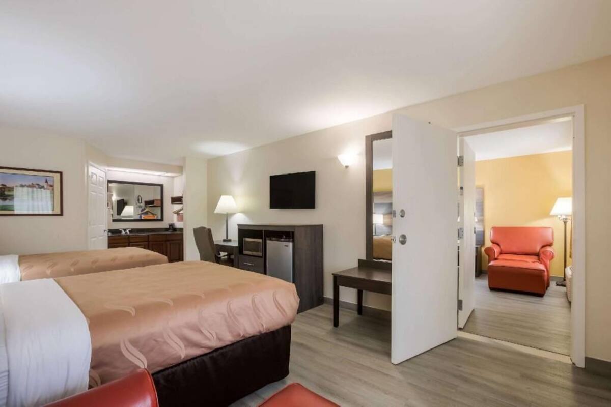 Ein Hotelzimmer mit zwei Betten, einem Schreibtisch, einem Stuhl, einem Fernseher und einer Tür zu einem weiteren Raum.