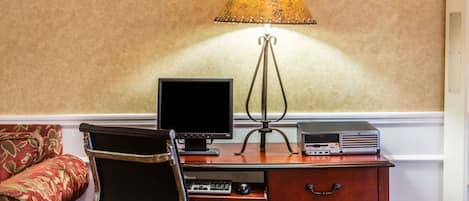 A wooden desk with a computer monitor, keyboard, and a lamp. There is a chair in front of the desk and a patterned sofa to the left.