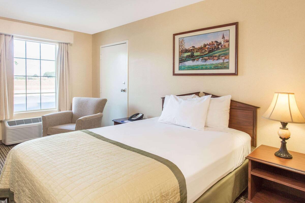A hotel room with a large bed, a chair, a bedside table with a lamp, a phone, and a painting on the wall.