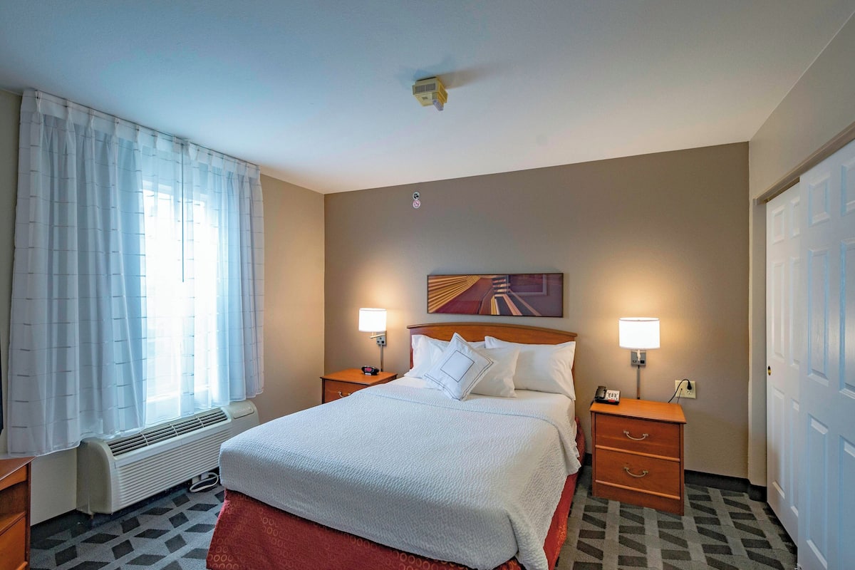 Habitación de hotel con una cama grande, dos mesitas de noche con lámparas, un radiador, una ventana con cortinas y una puerta.