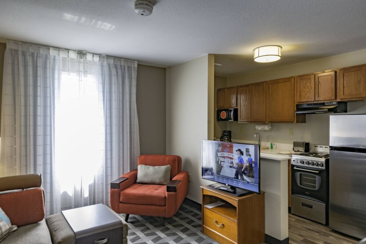 Habitación de hotel compacta con sofá, sillón, televisión, kitchenette y ventana con cortinas.