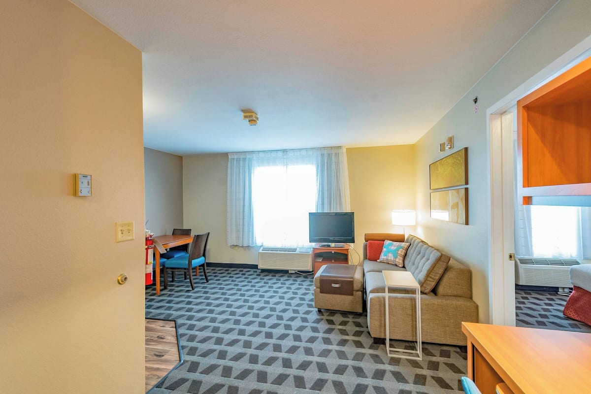 Habitación de hotel con sofá, televisión, una mesita, una silla y una ventana con cortinas.