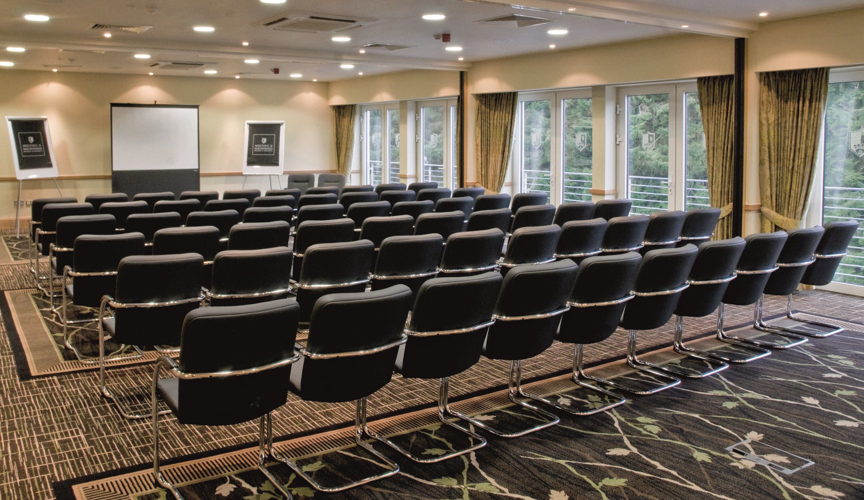 A conference room with rows of black chairs, a projector screen, and a banner on the wall.