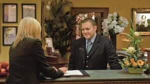 A reception desk with a man in a suit, a woman in a suit, and a vase of flowers.