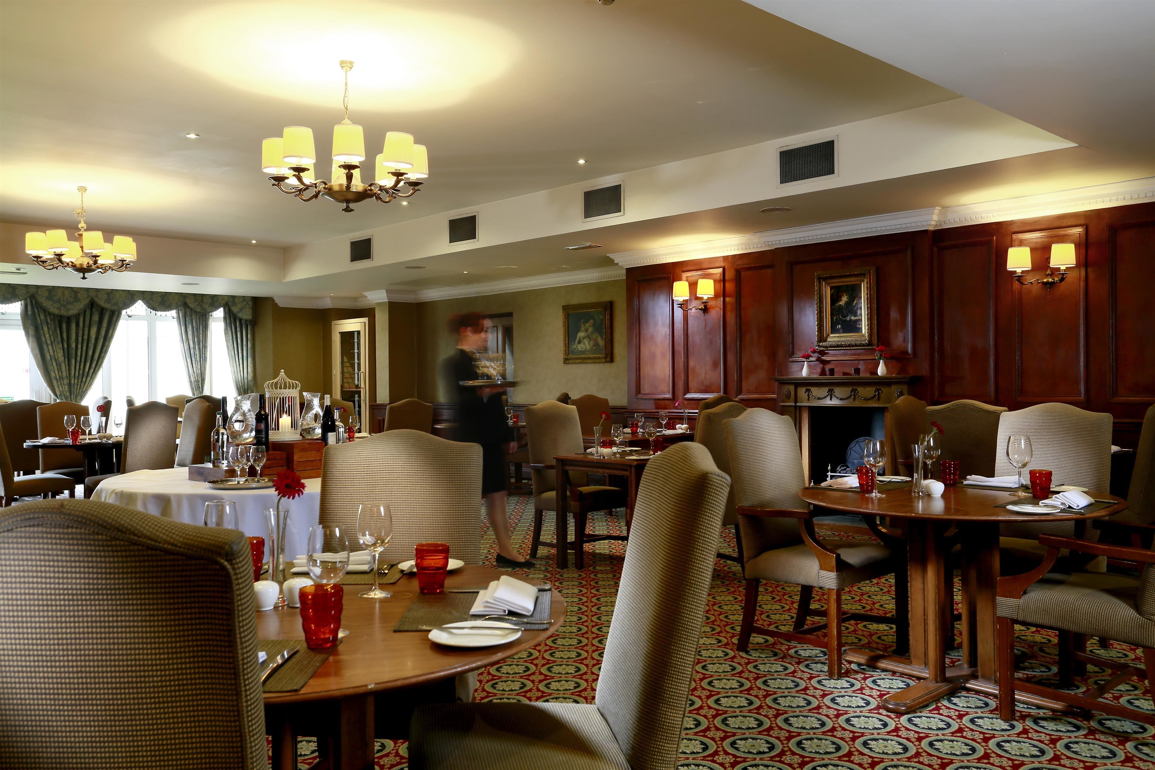 A hotel dining area with multiple tables and chairs, chandeliers, and a patterned carpet.