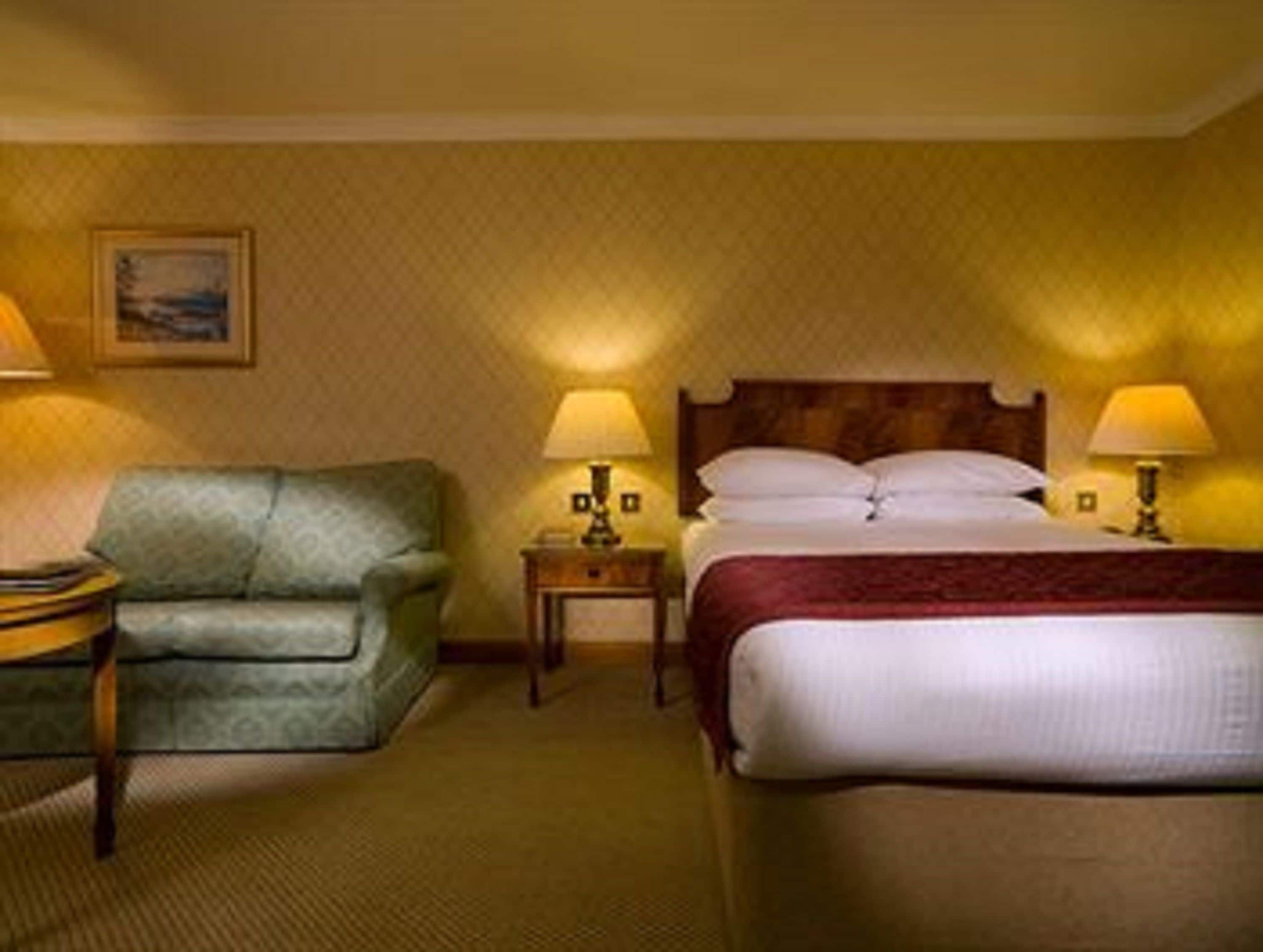 A hotel room with a bed, a green armchair, two bedside tables with lamps, and a painting on the wall.