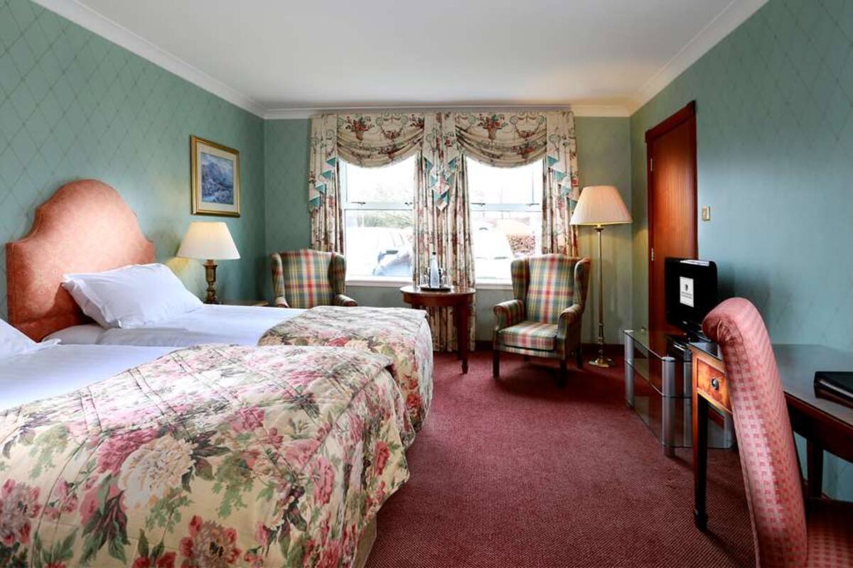 Ein Hotelzimmer mit zwei Betten, einem Schreibtisch, einem Stuhl, einem Fernseher und einem Fenster mit Vorhängen.