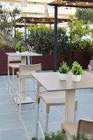 Espaço de pátio externo com mesas, cadeiras, plantas em vasos e uma estrutura de pérgola.