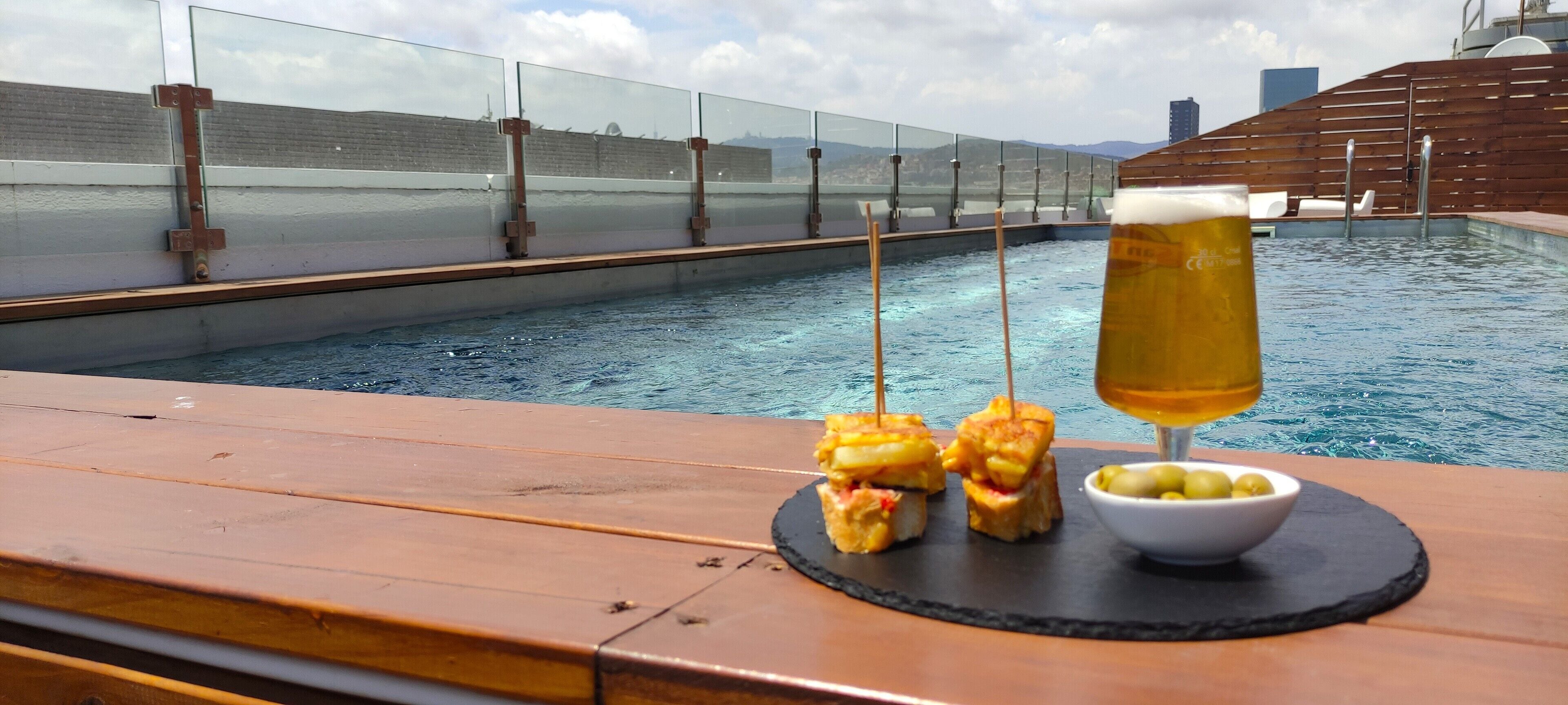 A poolside setting with a glass of beer, a bowl of olives, and grilled items on a wooden table.