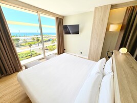 Une chambre d’hôtel avec un grand lit, un téléviseur à écran plat fixé au mur, une table de chevet avec une lampe, et une vue sur la ville par une grande fenêtre.