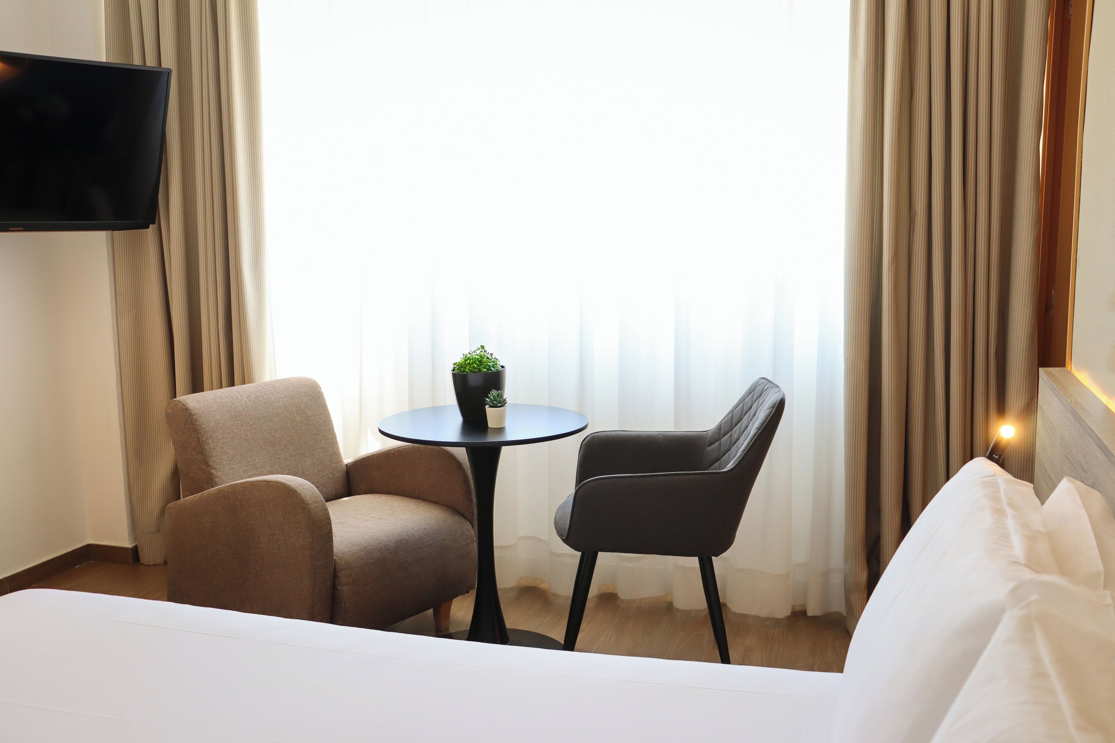A hotel room with a bed, two armchairs, a small table with a potted plant, a TV mounted on the wall, and a window with curtains.