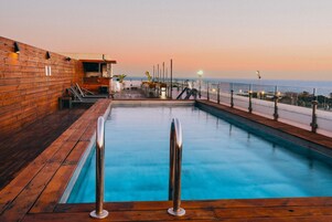 A pool area with wooden decking, metal railings, and a view of the ocean at sunset.