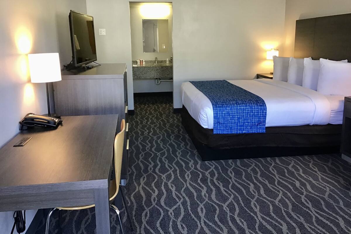 A hotel room with a bed, a desk, a chair, a TV, a mirror, and a bathroom visible through an open door.