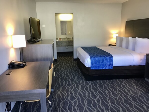 A hotel room with a bed, a desk, a chair, a TV, a mirror, and a bathroom visible through an open door.