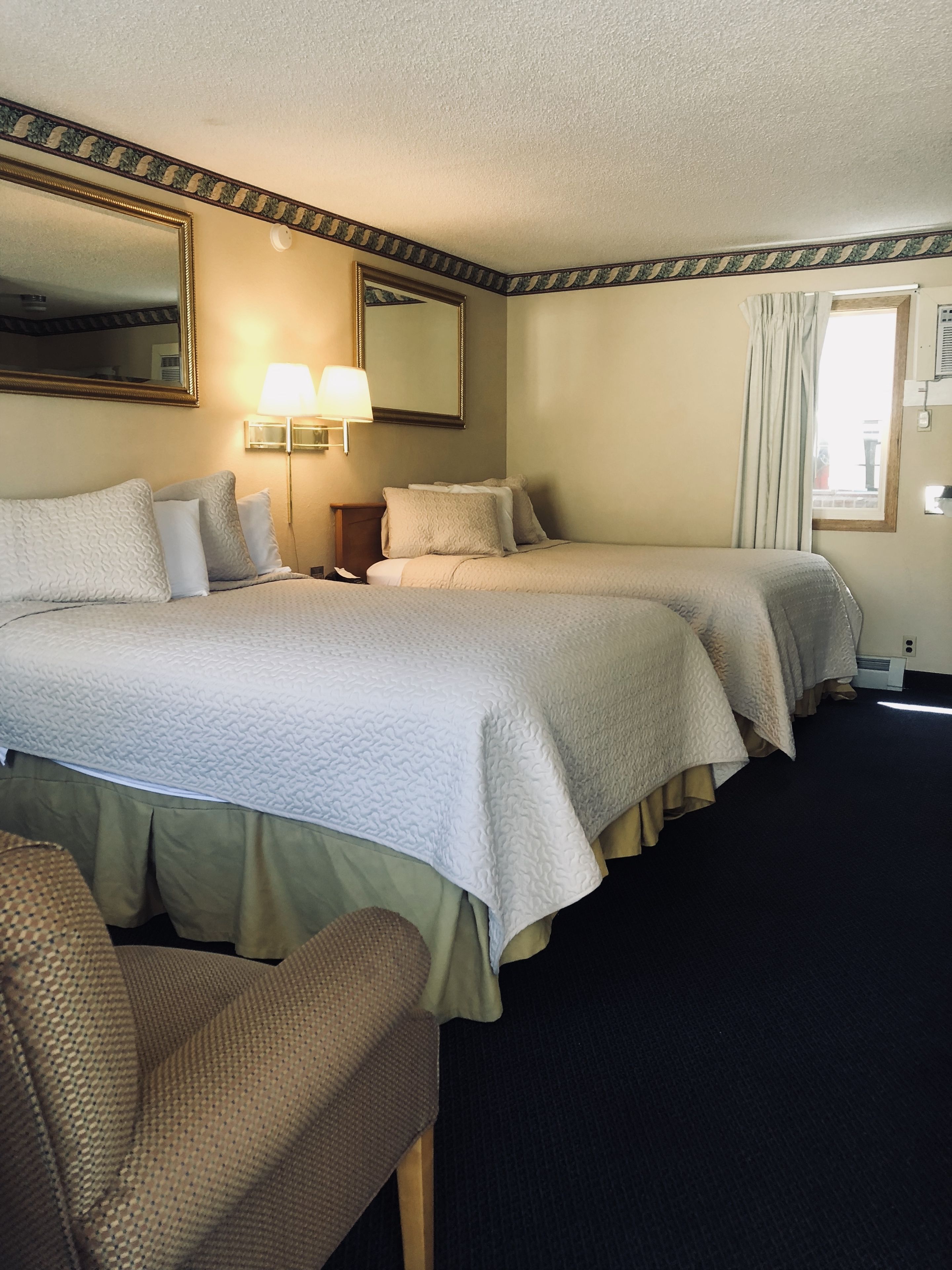 A hotel room with two beds, a nightstand, a lamp, a mirror, and a window with curtains.