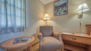 A hotel room with a chair, a small table with a magazine, a lamp, a painting on the wall, and a wooden dresser.