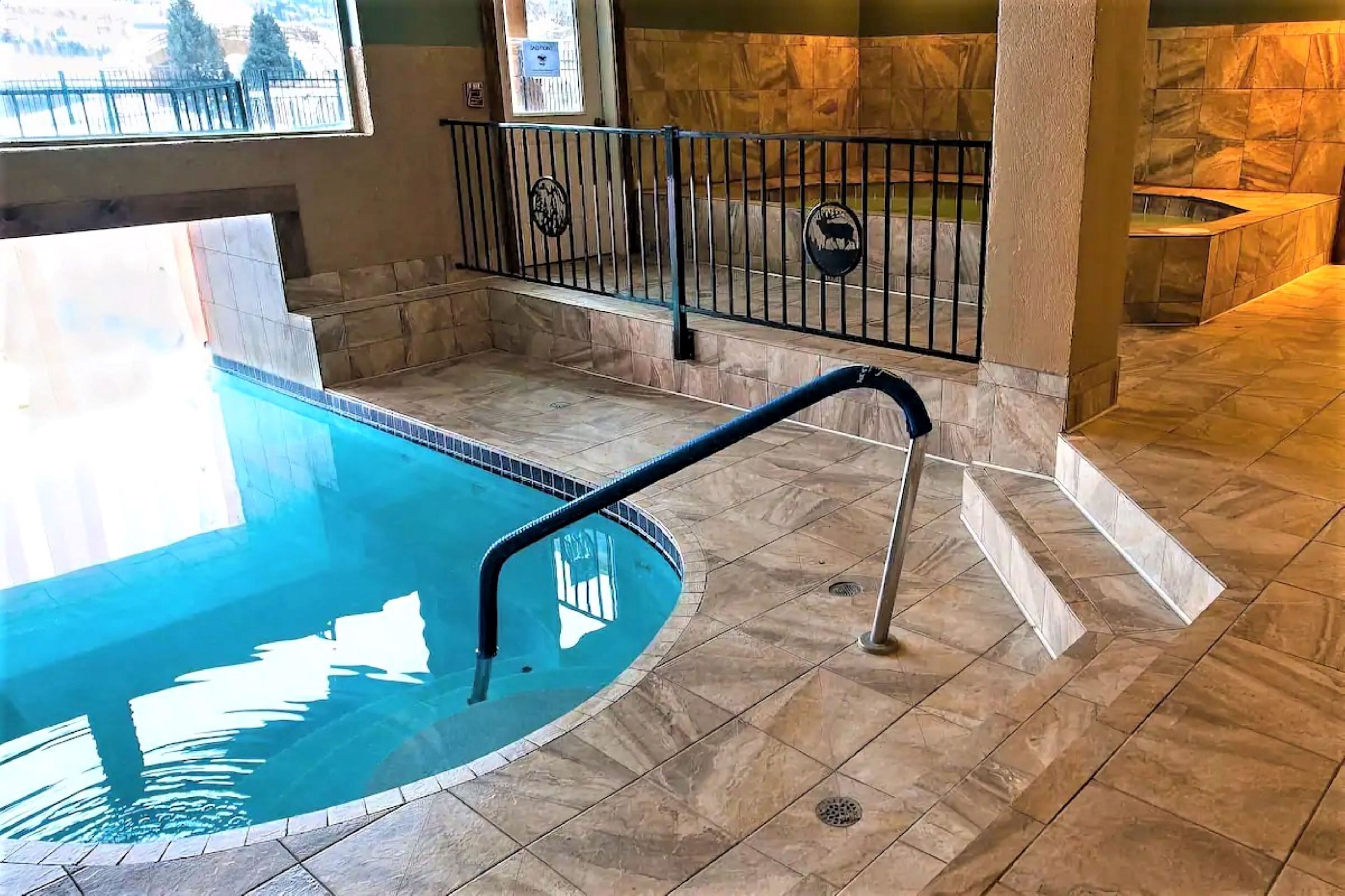 Una piscina interior con suelo de baldosas, barandilla metálica y pared de piedra al fondo.